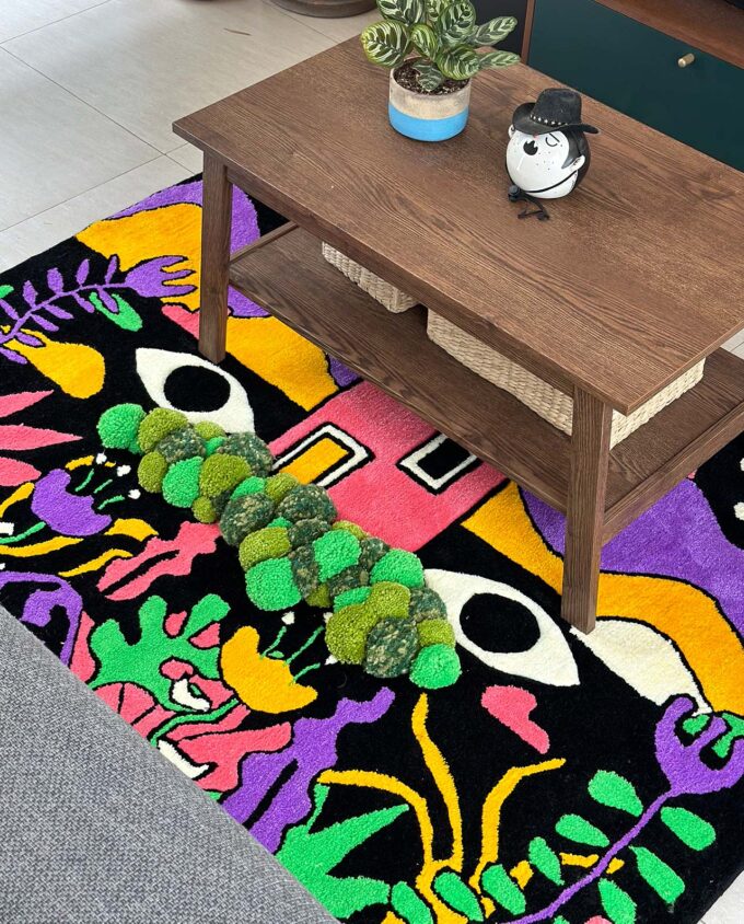 Detalle de una sección del tapete mundo cósmico hecho con técnica de tufting en México, con una casa al centro y unos ojos a los lados, rodeado de plantas con colores psicodélicos, abajo de una mesa de centro