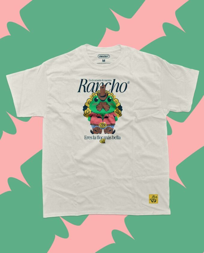 Playera color beige con la ilustración de una rana ranchera verde con un ramo de flores amarillas, y una frase que dice: En el corazón de nuestro rancho, eres la flor más bella