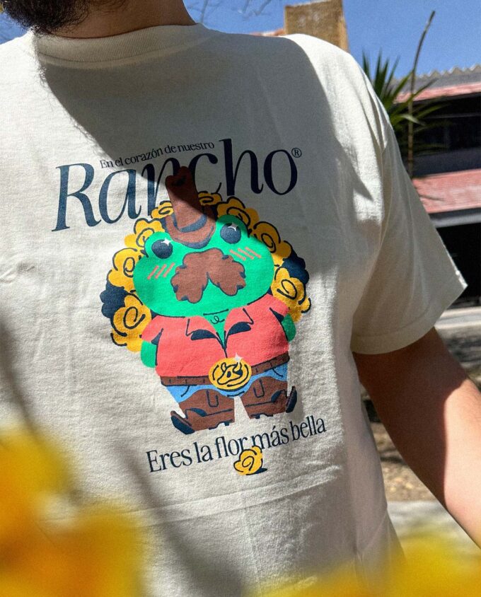 Fotografía de detalle de una playera color beige con la ilustración de una rana ranchera verde con un ramo de flores amarillas, y una frase que dice: En el corazón de nuestro rancho, eres la flor más bella