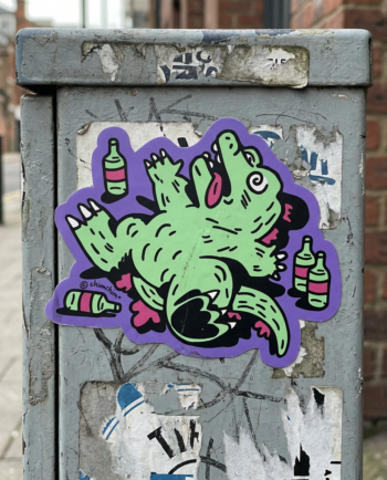Sticker vinil matte Drunkzilla 9x7.5 cm, lagarto verde patas arriba rodeado de botellas sobre fondo morado, contorno recortado ChimChim México